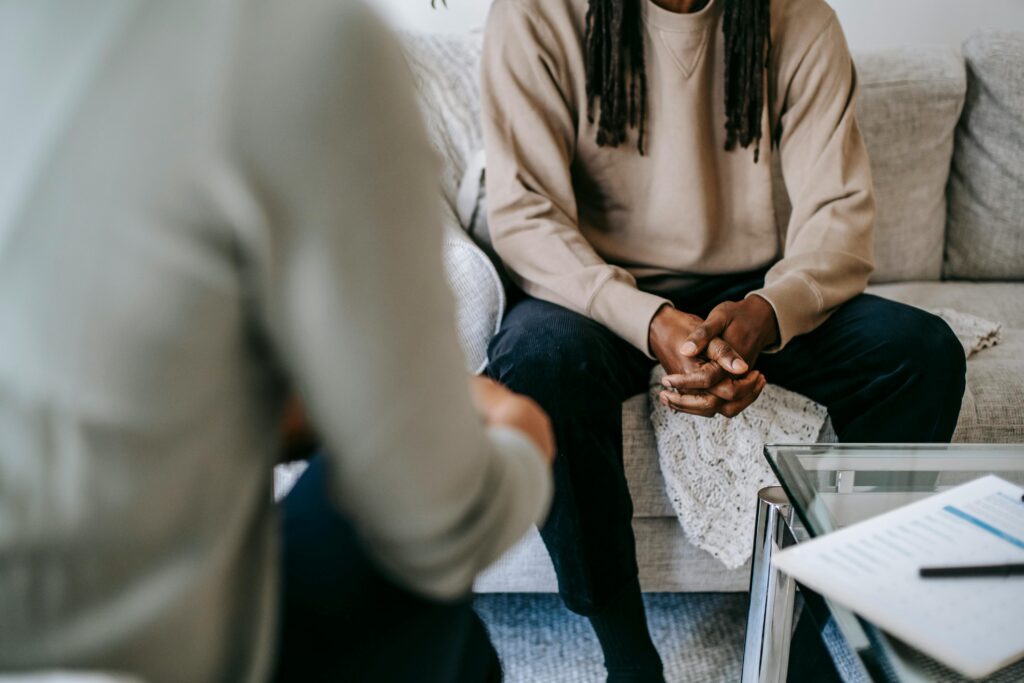 Unrecognizable African American male patient with clasped hands sitting on sofa near blurred anonymous therapist during visit in psychotherapy office